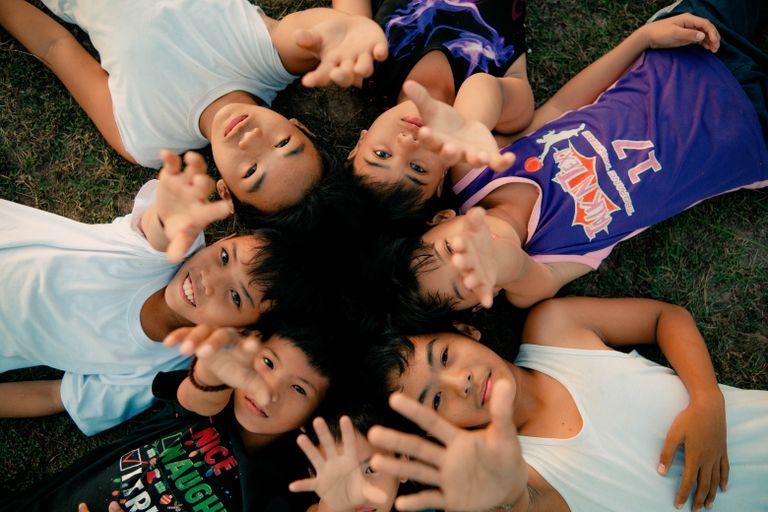 A group of Filipino children lying on the grass, reaching towards the camera with smiles.