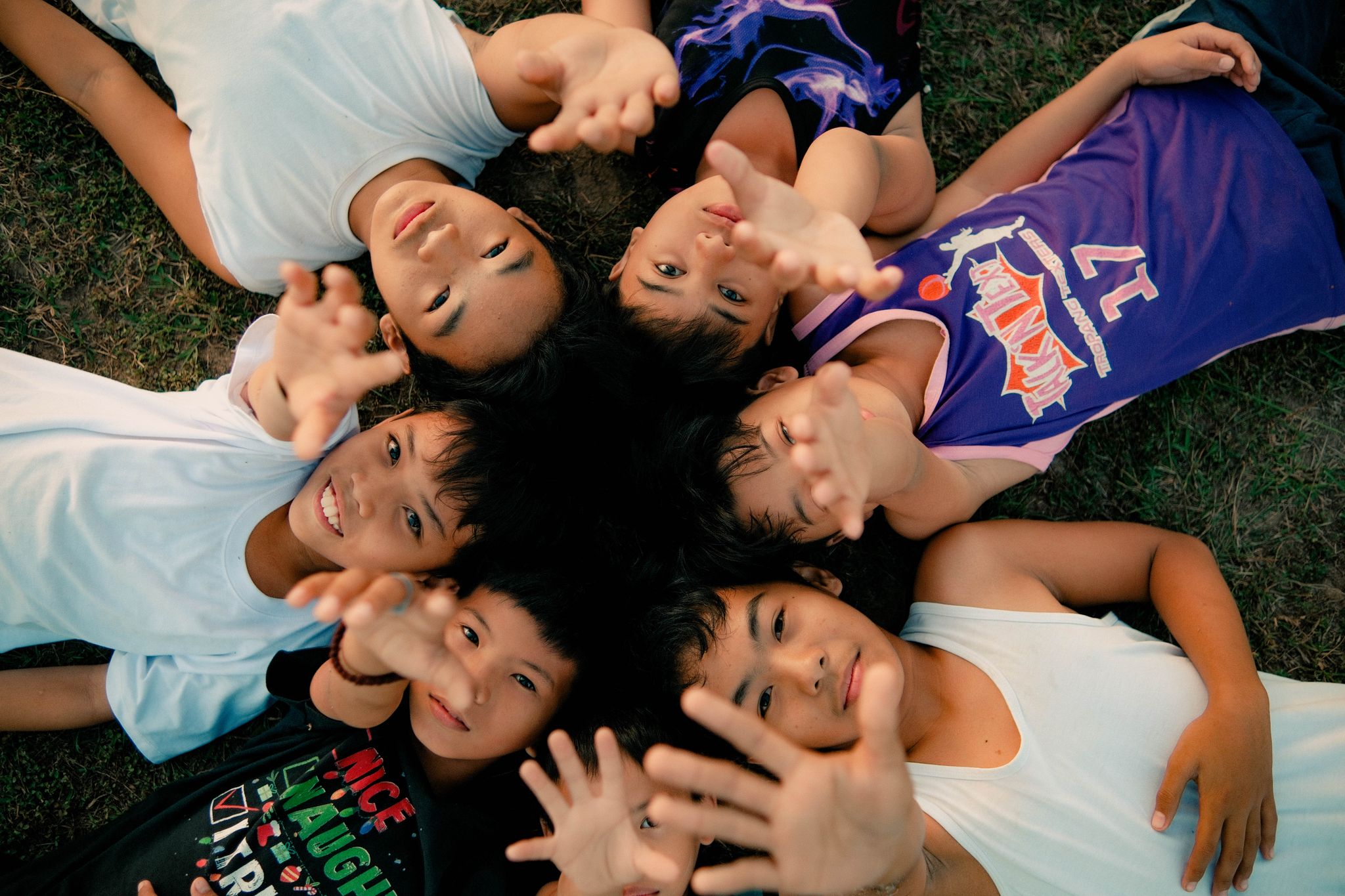 A group of Filipino children lying on the grass, reaching towards the camera with smiles.