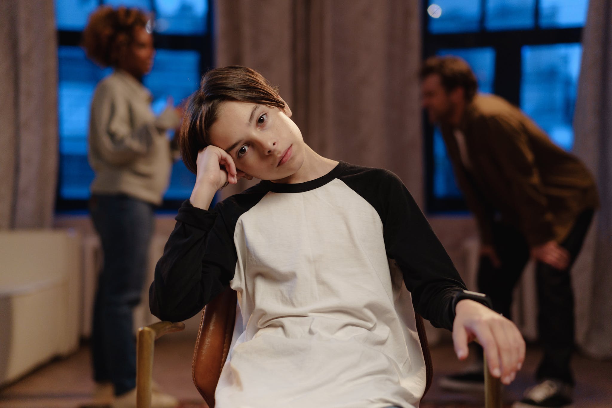 A young boy in a black and white shirt looks sad as his parents argue in the background.