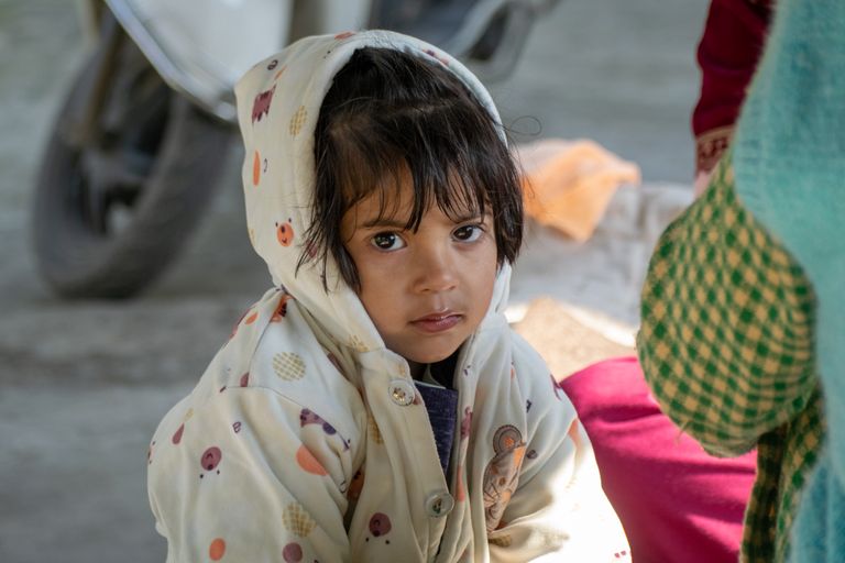 A young child sitting indoors, wearing a patterned hoodie, looking directly at the camera.