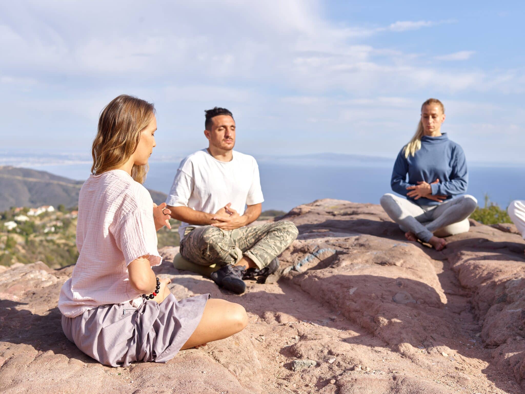 Group of three individuals practicing meditation on a rocky outcrop with a scenic view of hills and the ocean in the background. The setting promotes mindfulness and tranquility.