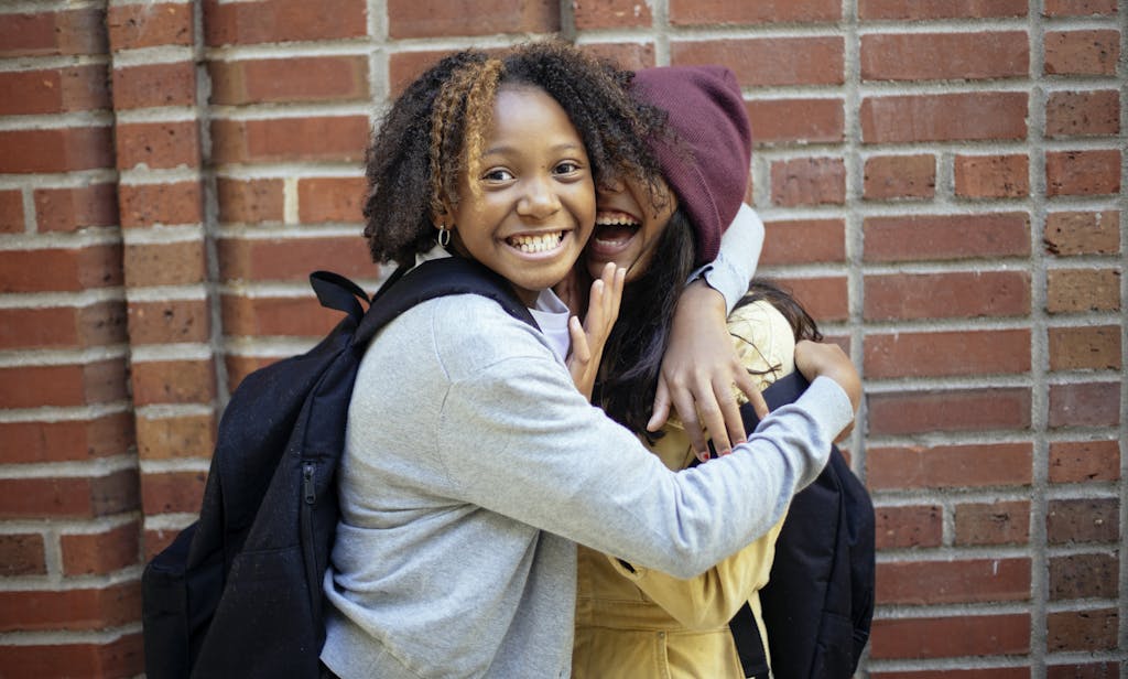 Cheerful African American girl in casual clothes hugging friend while having fun near brick wall