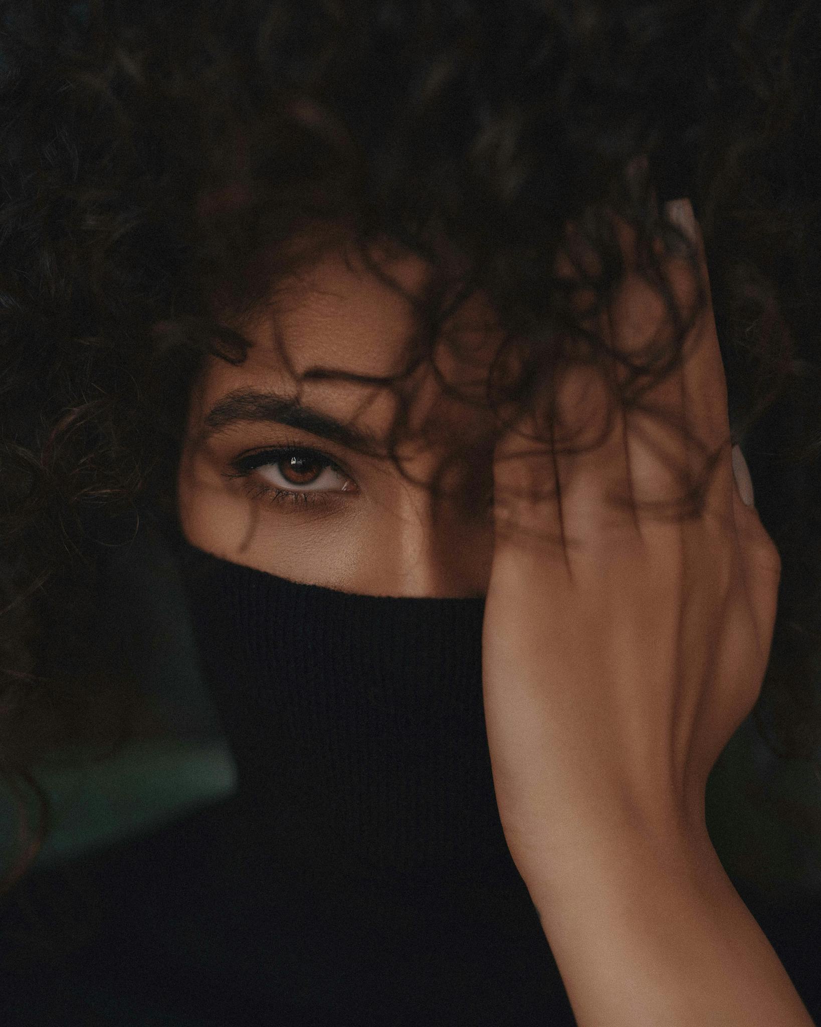 Close-up of a woman with curly hair, partially hiding her face with a black turtleneck and hand.