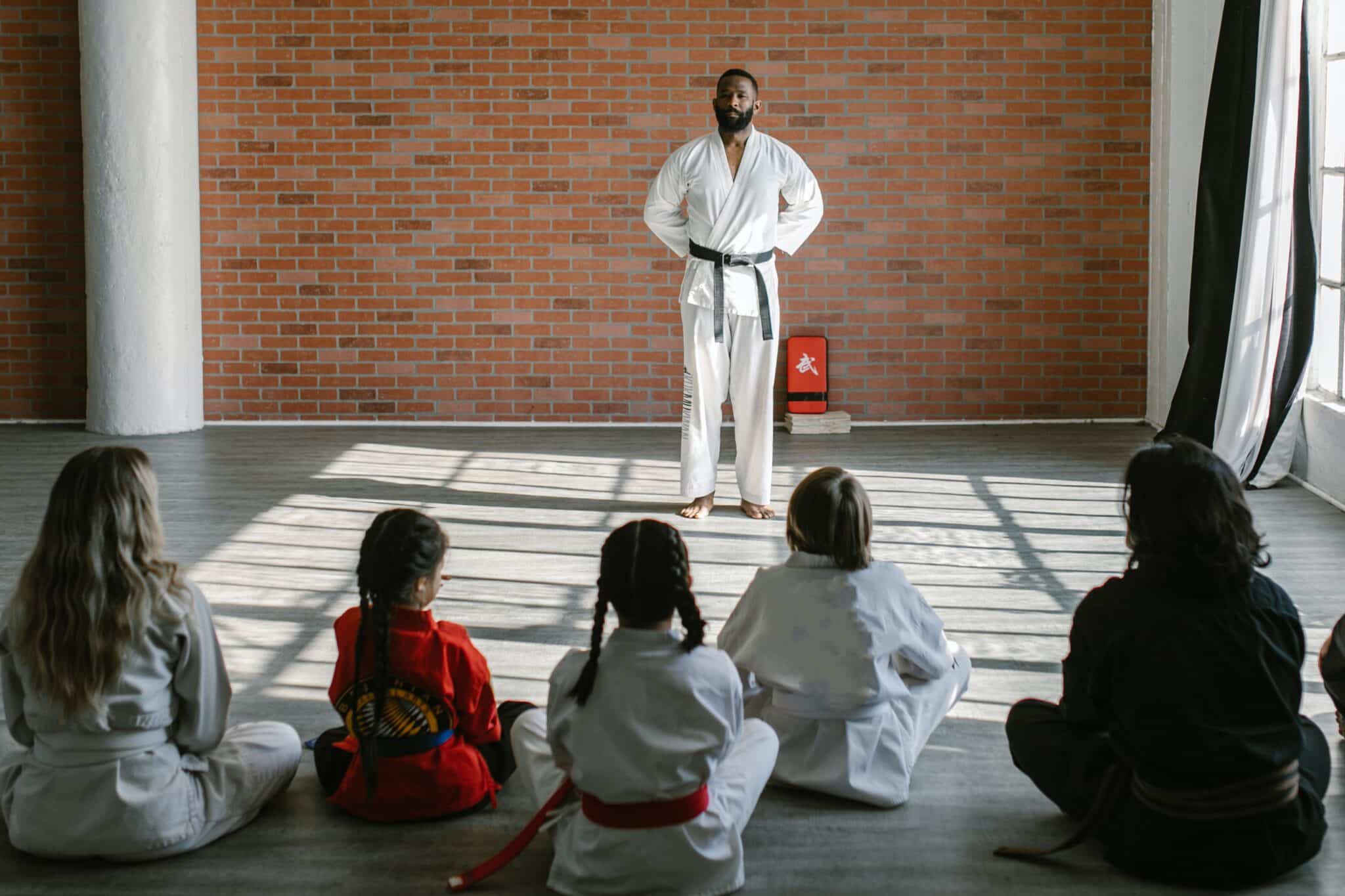 Martial arts instructor in white gi with black belt teaching students sitting on floor in dojo with brick wall.