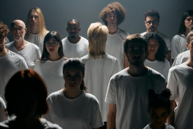 A diverse group of individuals of various ages and ethnicities standing together in a dimly lit space, all wearing plain white t-shirts, creating a unified yet distinct visual representation of community and individuality.