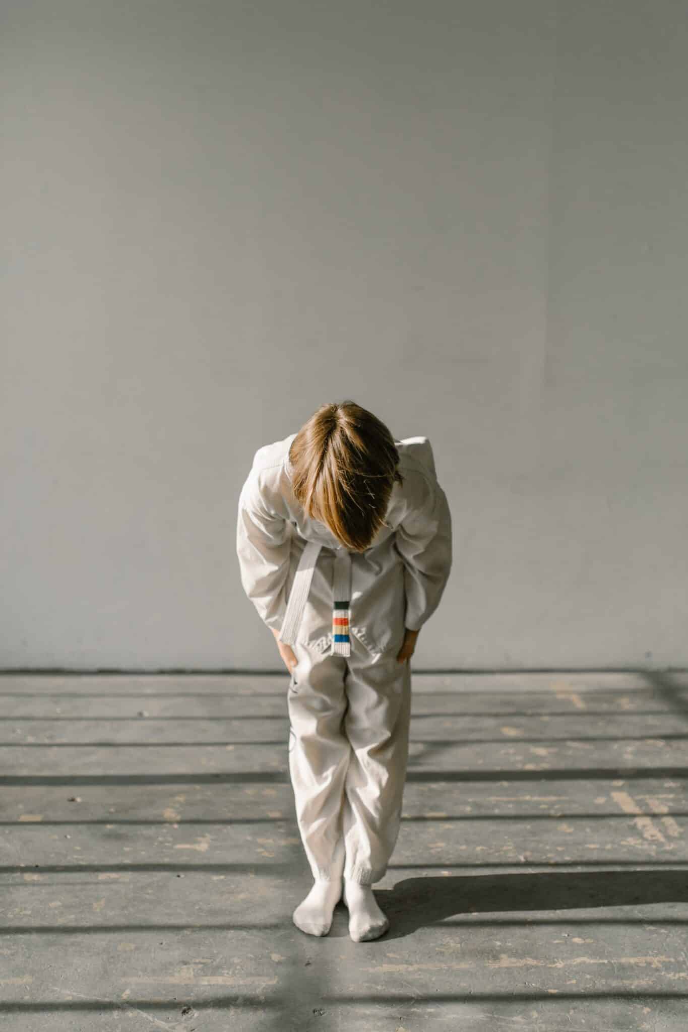 Young child in martial arts uniform bowing on concrete floor with shadows.