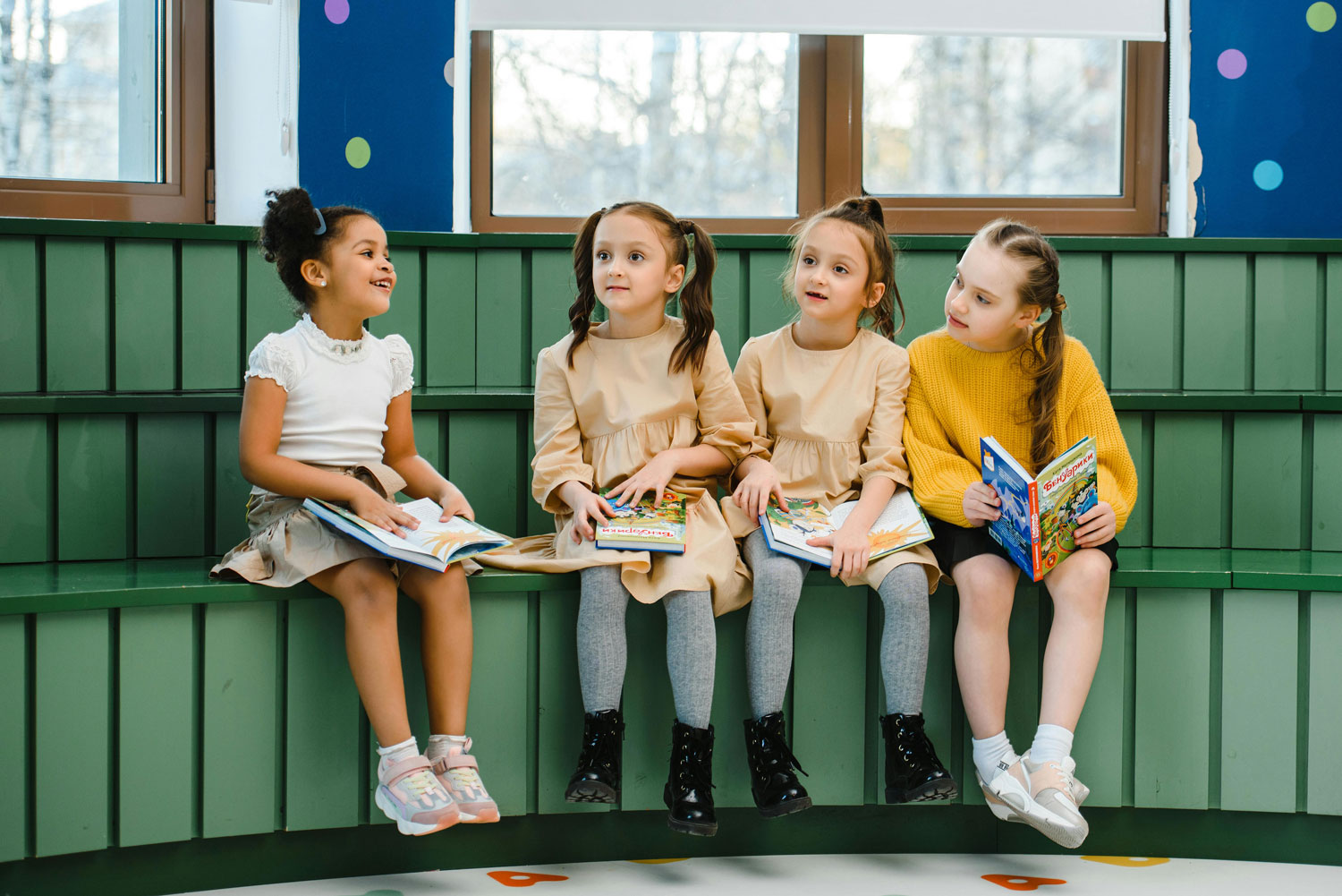 Four young girls sitting on a green bench by windows, holding and reading children's books in a colorful classroom.
