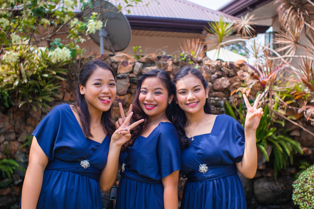 Three young women in blue dresses smiling and posing outdoors as bridesmaids, showing peace signs.
